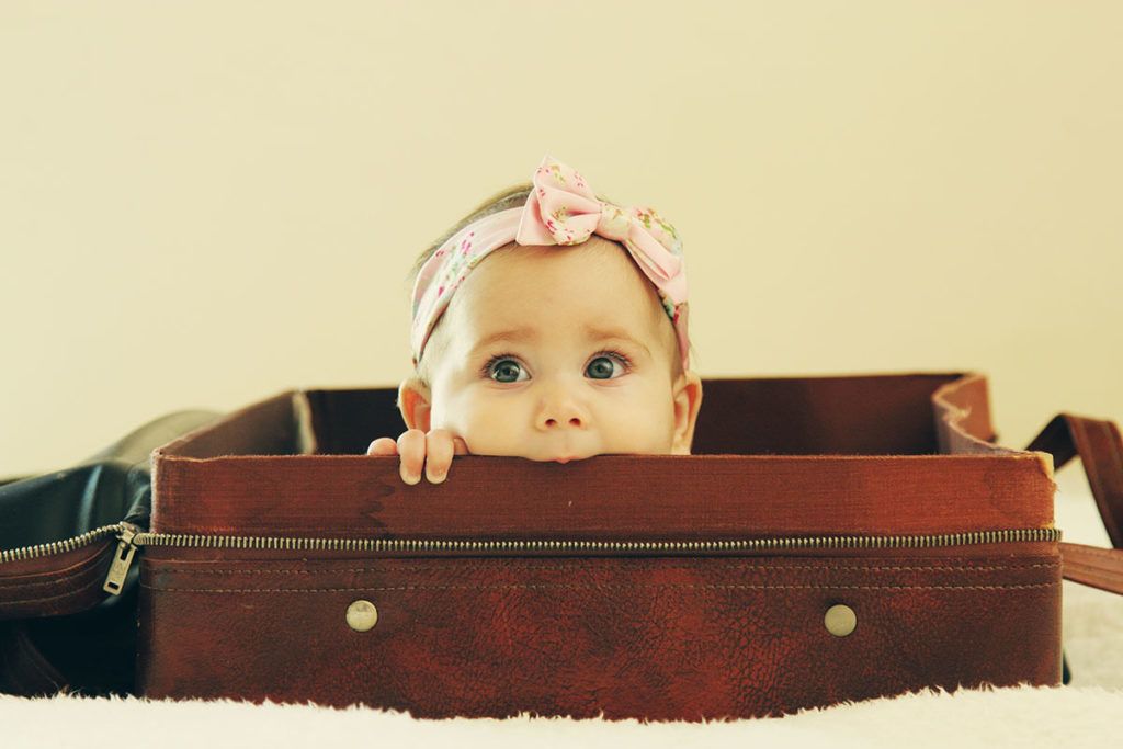 Ein kleines Baby mit Schleifchen auf dem Kopf liegt in einem braunen Koffer und schaut über den Rand und knabbert dabei am Koffer.