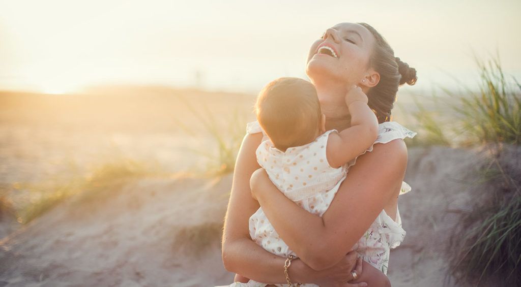 Eine Mutter sitzt mit ihrem Baby auf dem Arm im Sand, das Baby zwickt ihr in den Hals, sie lacht.