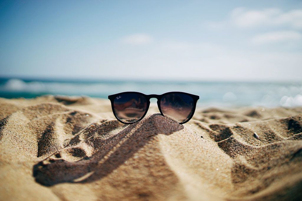 Eine schwarze Sonnenbrille liegt auf einem kleinen Sandhaufen am Strand. Im Hintergrund glitzert das Meer.