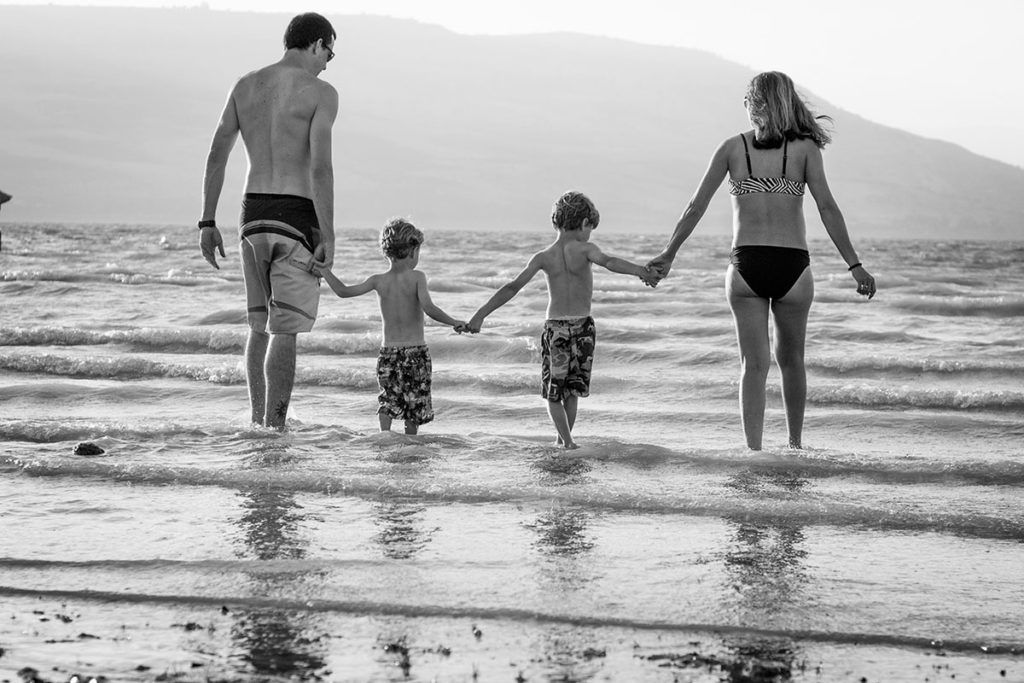 Eine junge Familie (Vater, zwei Söhne und Mutter) laufen in Badekleidung Hand in Hand in das Meer hinein. Das Bild ist schwarzweiß.