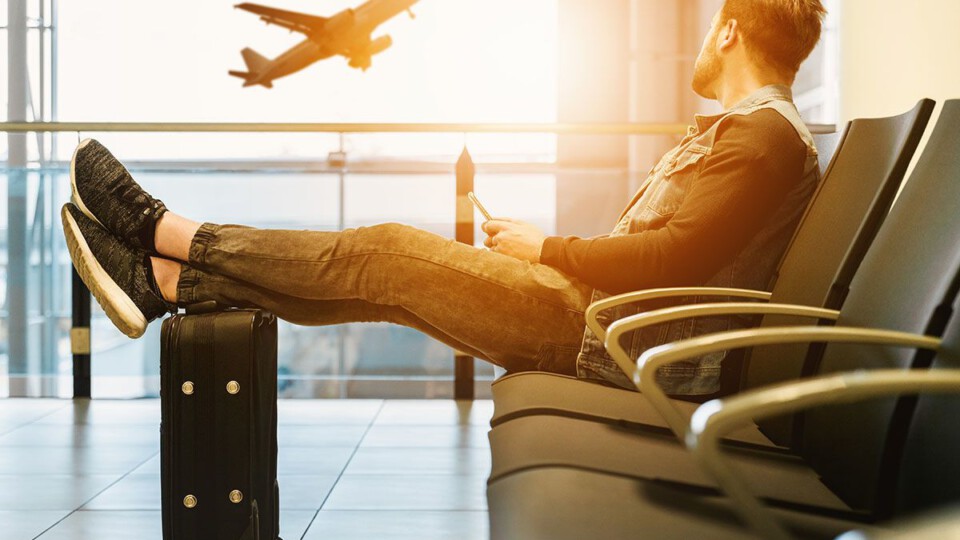 Ein Mann sitzt am Flughafen Terminal, hat die Füße auf seinem Koffer gelegt und sieht aus dem Fenster. Im Hintergrund startet ein Flugzeug in die Sonne.
