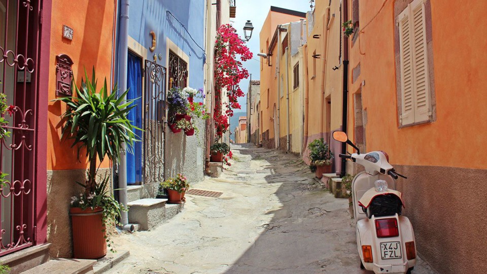 Eine enge Gasse in Italien, die Häuser rechts und links sind bunt angestrichen, Topfpflanzen verzieren die Fassaden, eine weiße Vespa steht rechts unten